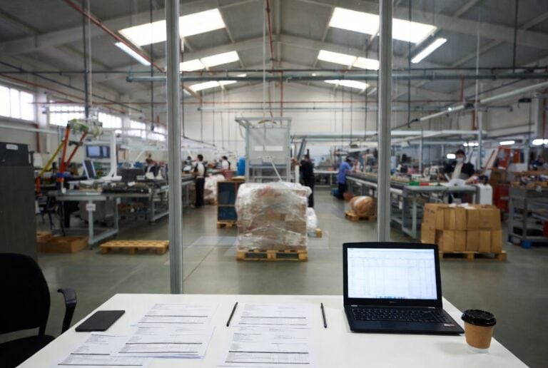 Factoring per le PMI: scrivania con documenti e laptop in primo piano, operai al lavoro nella linea produttiva oltre il vetro